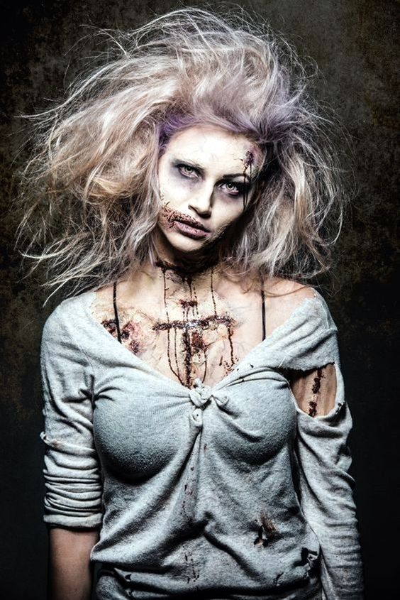 This zombie look features wild, teased hair, pale skin with dark, sunken eyes, and stitchedup wounds on the face and chest. The tattered, distressed shirt completes the creepy, undead appearance.