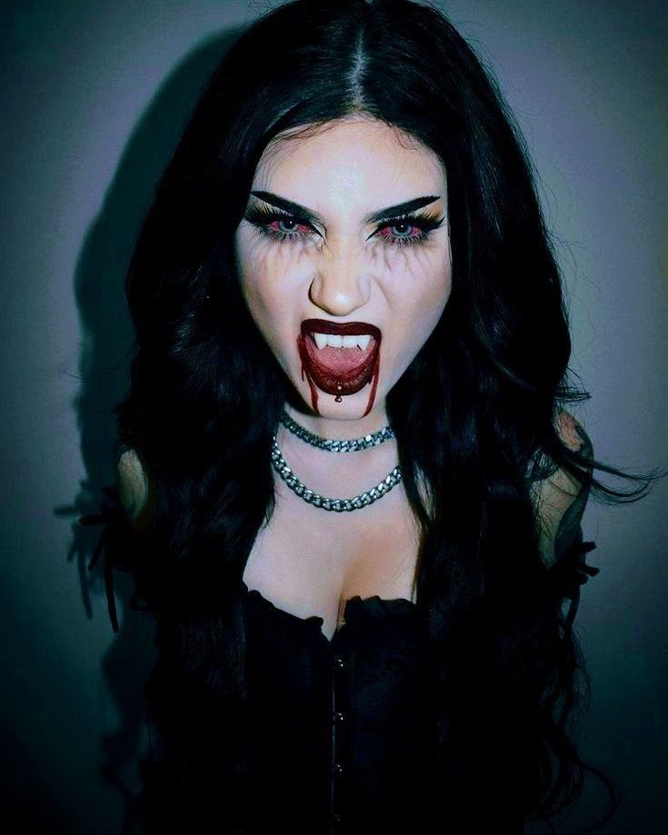 This vampire Halloween look features dark, dramatic eye makeup, blood-red lipstick with drips extending down the chin, and fangs, creating a fierce, intense expression. The dark hair and accessories add to the classic, spooky vampire vibe.