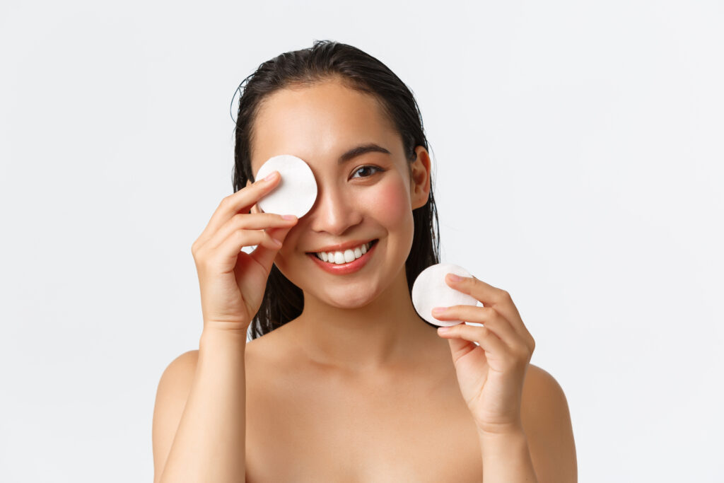 Skincare, women beauty, hygiene and personal care concept. Close-up of attractive naked asian woman in bathroom, wipe-off makeup with cotton pads and smiling camera, white background.